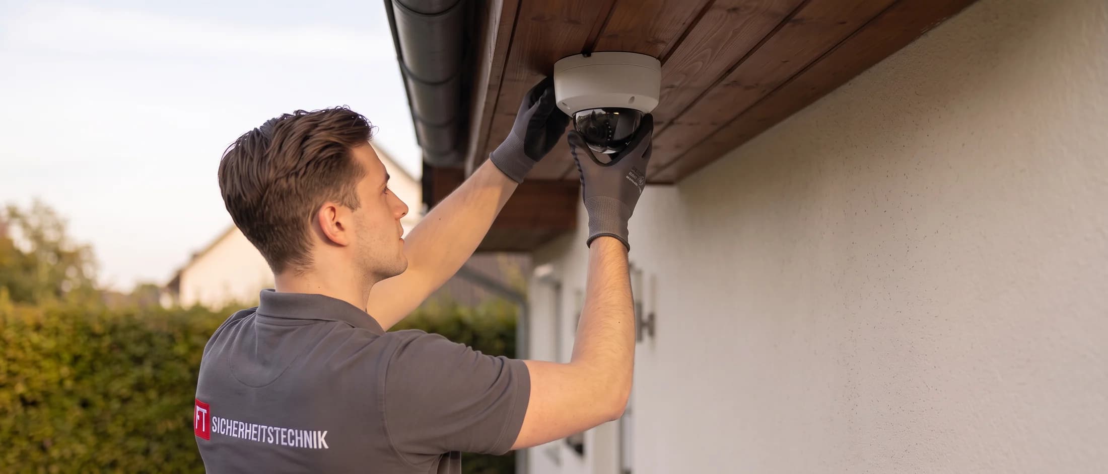 Techniker der FT Sicherheitstechnik setzt eine Dome-Kamera in die Holzverkleidung des Dachüberstands ein, die Zuleitung liegt unsichtbar dahinter